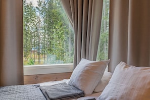 A bedroom with a bed, pillows, a quilted blanket, and a view of trees through a window with curtains. - Saimaa Life & Light (Imatra)