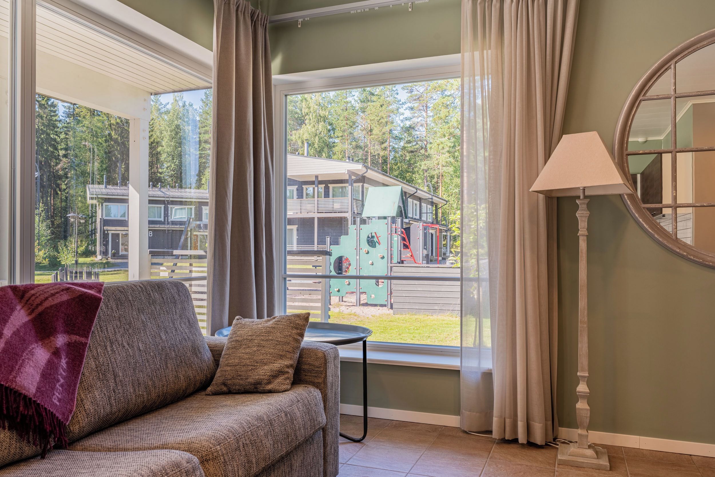 A room with a sofa, a lamp, and a view of a playground and buildings outside.