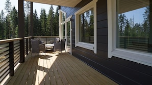 A wooden deck with outdoor furniture, large windows, and a view of a forest.