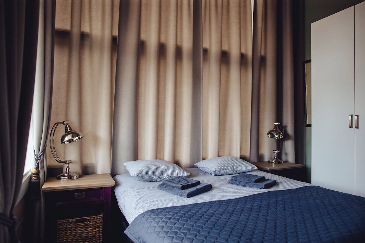 Cama bem arrumada com colcha azul, dois travesseiros e duas luminárias laterais em uma mesa de cabeceira.