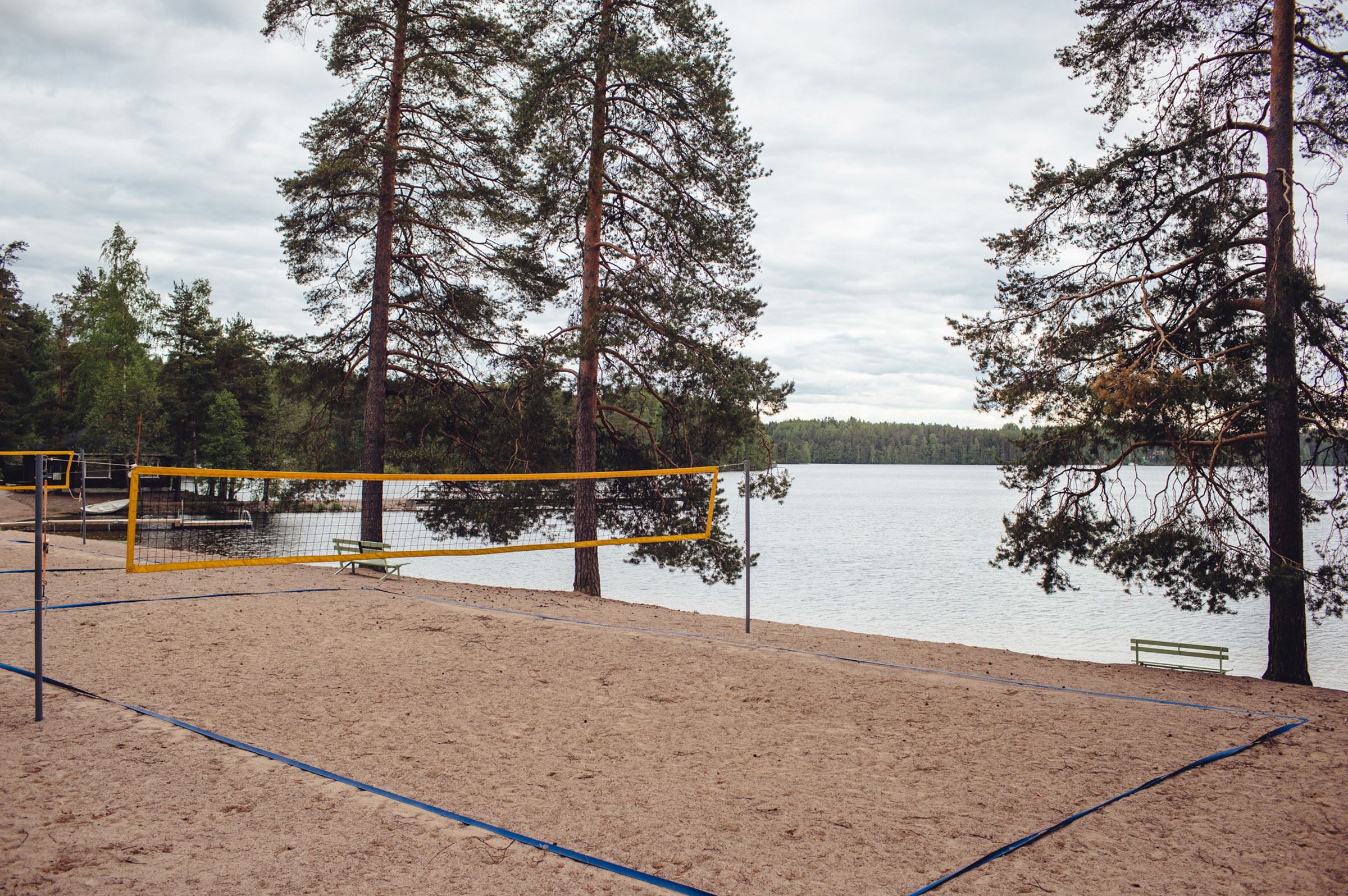 Una cancha de voleibol de playa con red, rodeada de arena y árboles, con vista a un lago tranquilo.