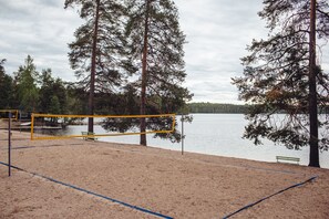 Een strandvolleybalveld met een net, omringd door zand en bomen, met uitzicht op een kalm meer.
