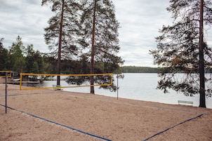 Una cancha de voleibol de playa con red, rodeada de arena y árboles, con vista a un lago tranquilo.