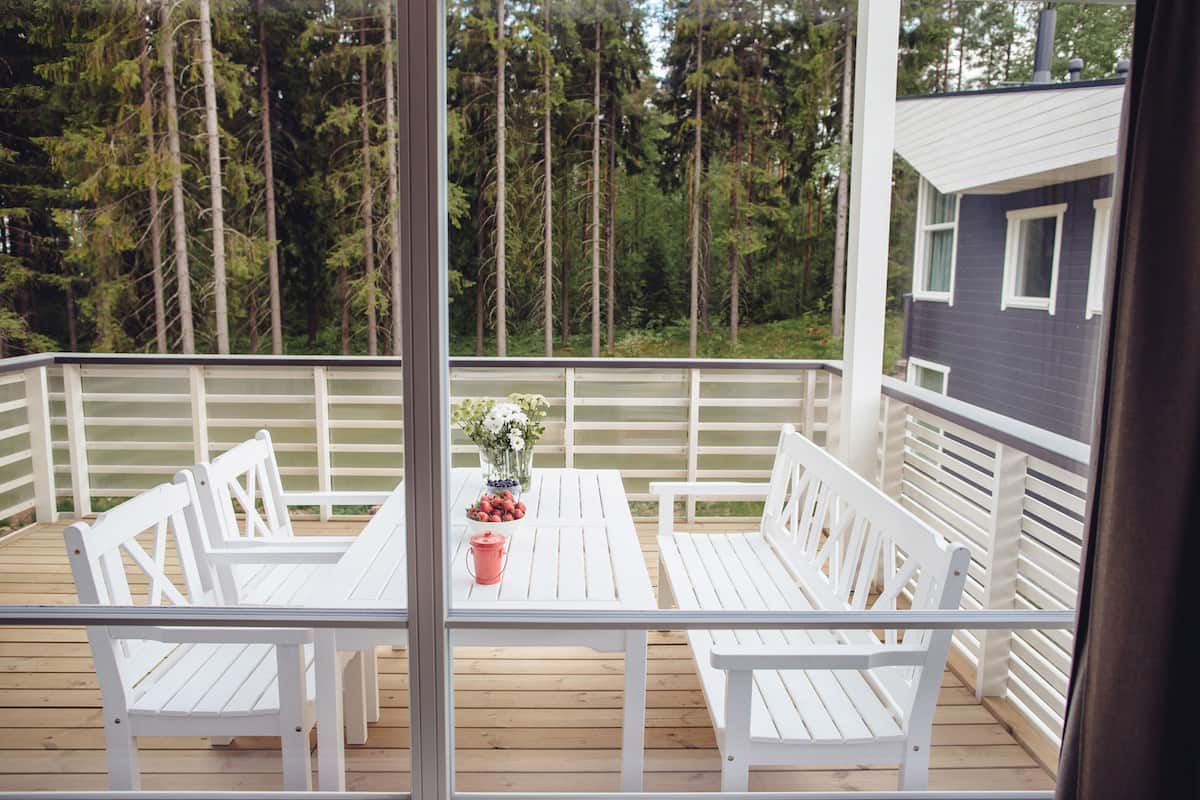 Um deck de madeira com móveis brancos, uma mesa com um vaso de flores e vista para uma área arborizada.