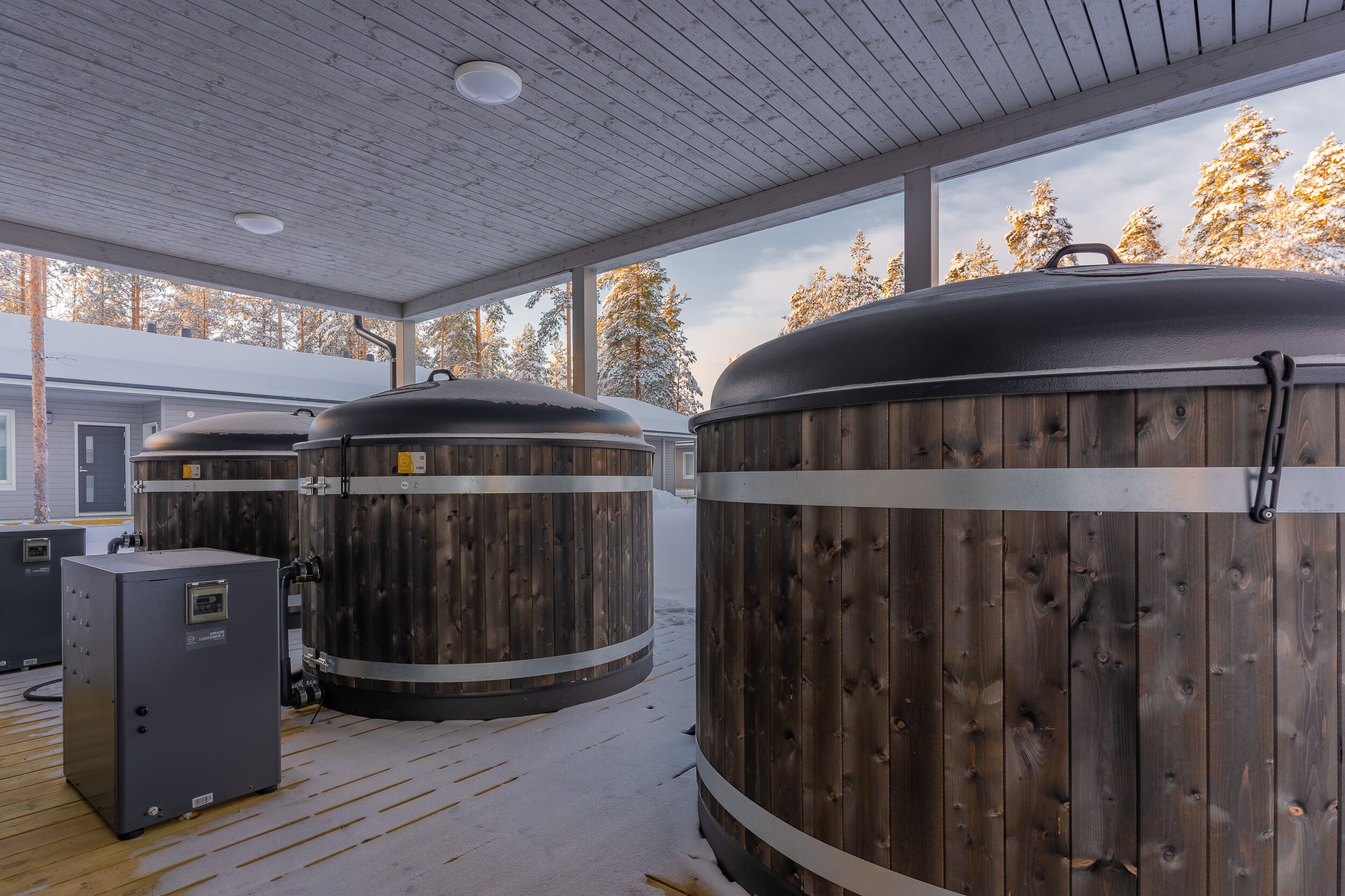 Un espacio exterior cubierto con tanques cilíndricos grandes, piso de madera y vistas a árboles nevados a través de amplias ventanas.