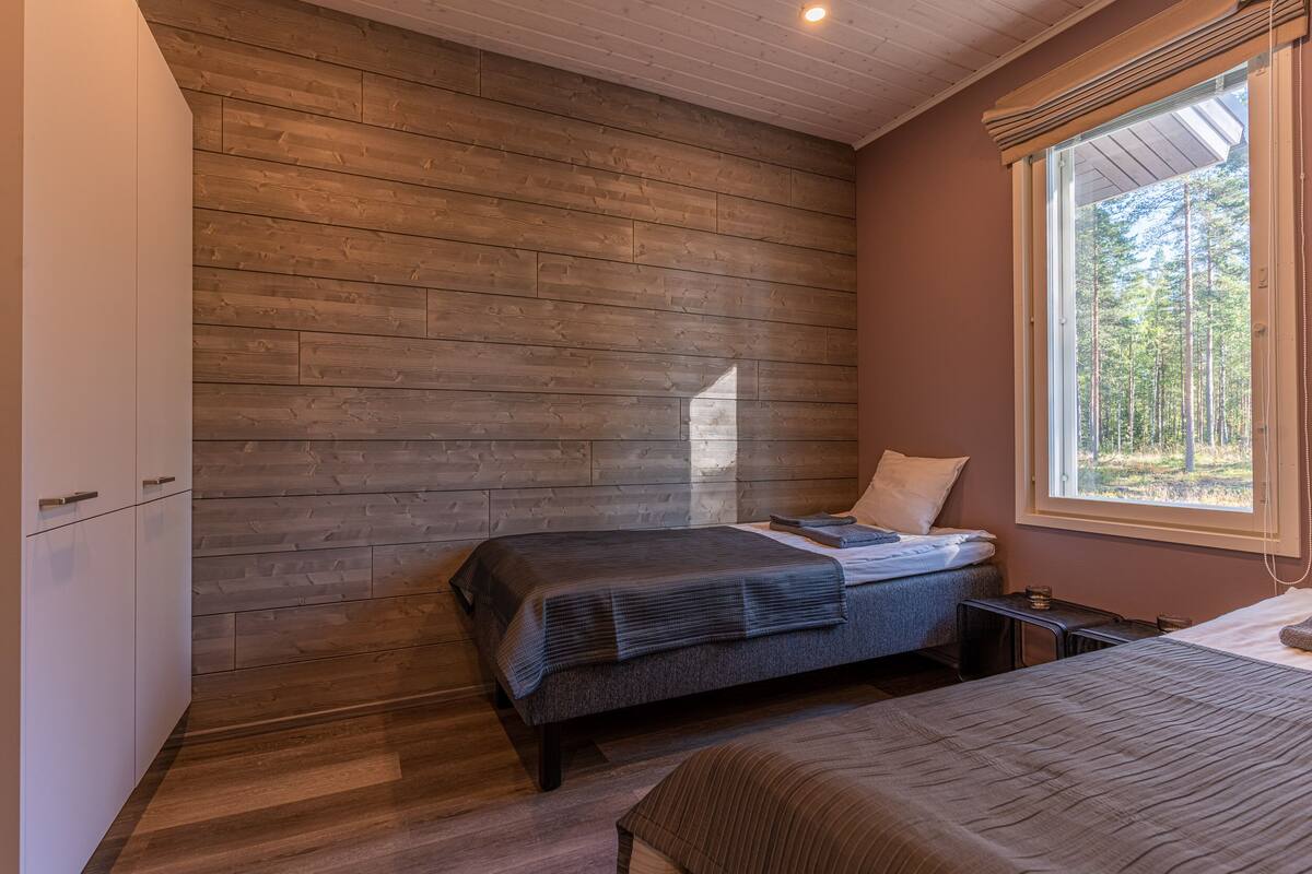Quarto com parede de madeira, uma cama, janela com vista para árvores e uma pequena mesa de cabeceira.