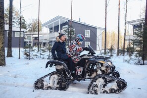 Een sneeuwscooter met een bestuurder in een geruit jasje en helm, geparkeerd in een besneeuwd gebied met huizen op de achtergrond.