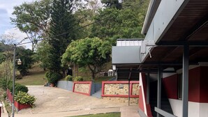 A building with a red and grey facade, a stone pathway, and a grassy area with trees. - Monique's Guesthouse (Port of Spain)
