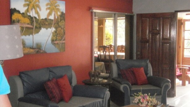 A living room with two grey sofas, red cushions, a wooden coffee table, and a painting of palm trees on the wall. - Monique's Guesthouse (Port of Spain)