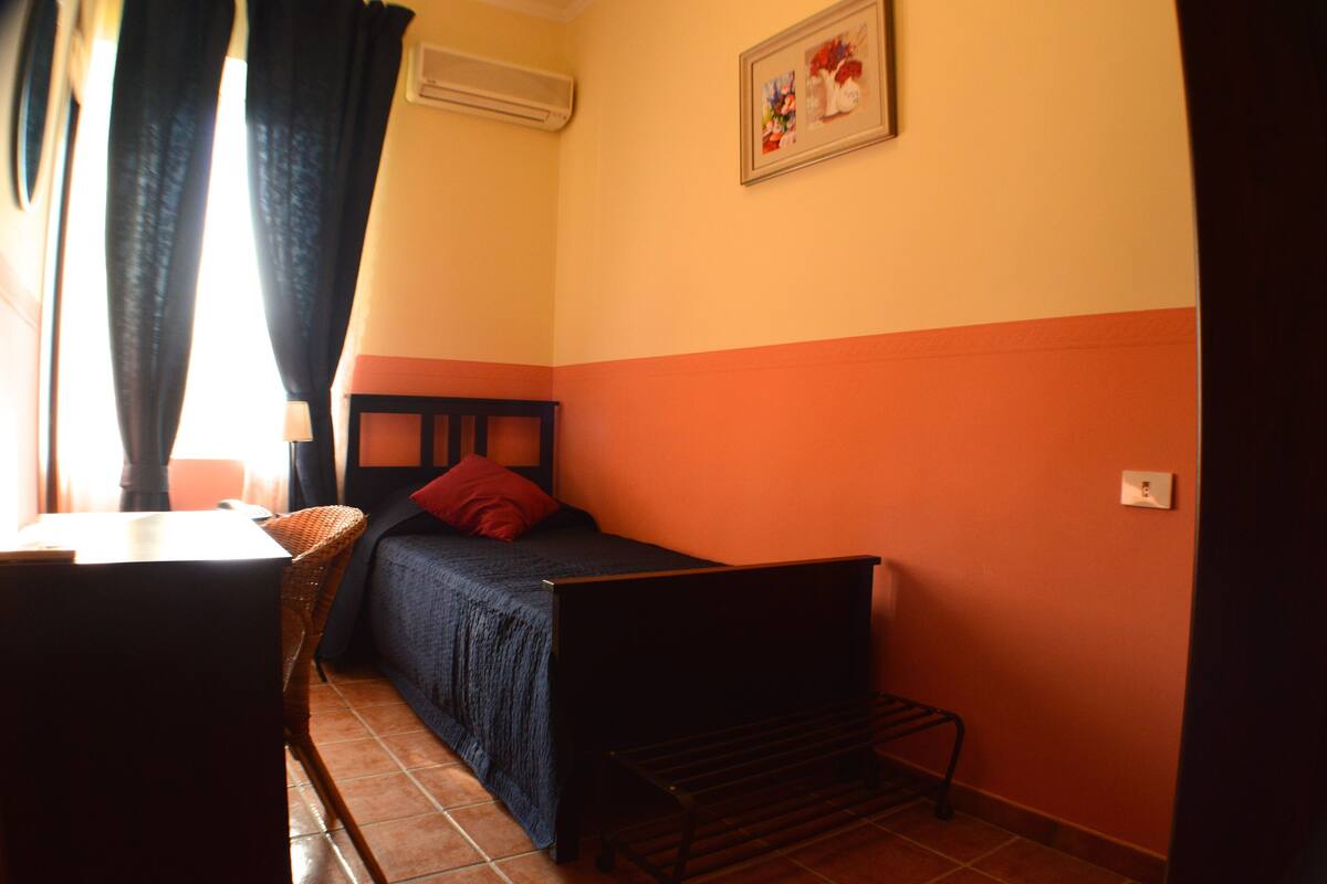 A small, single-bed room with a wooden headboard, a red pillow, and a chair.
