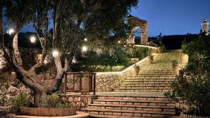 A stone staircase with a wooden railing, surrounded by potted plants and trees, leading up to a stone structure with a gate. - Nobelos Seaside Lodge (Zakynthos)