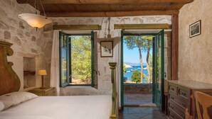 A bedroom with a wooden bed, a nightstand with a lamp, a dresser, and a view of the sea through open doors. - Nobelos Seaside Lodge (Zakynthos)