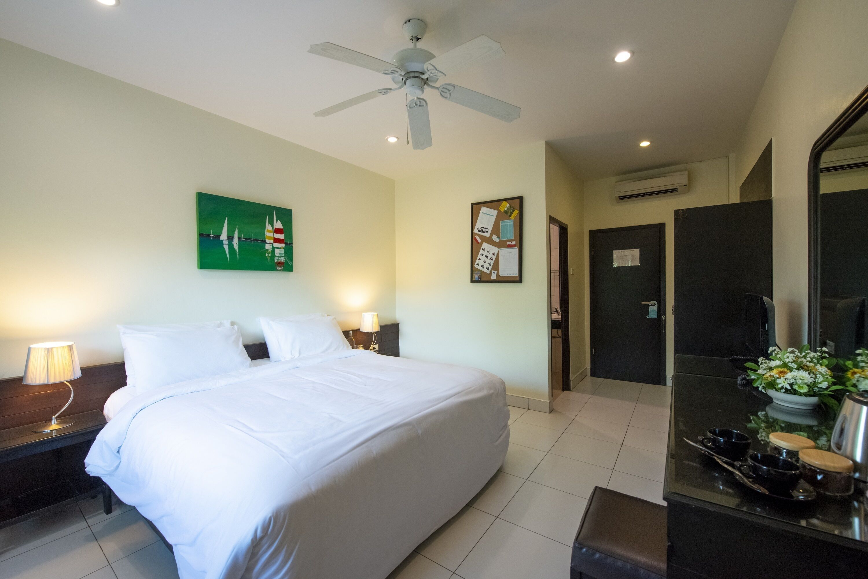 Habitación de hotel con una cama grande, un ventilador de techo, un cuadro de veleros, un tablón de anuncios y una mesita con un florero.