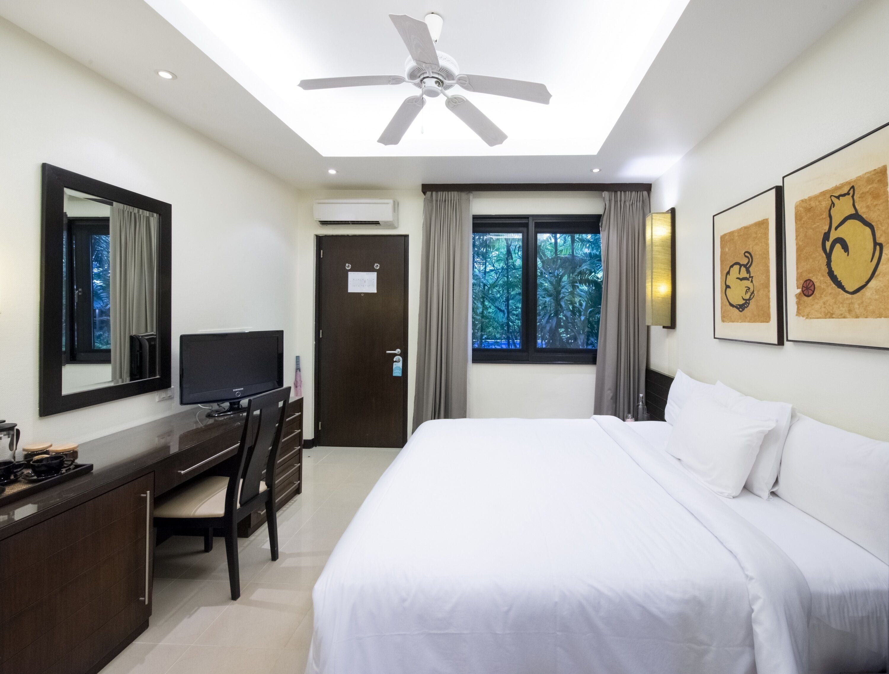 A hotel room with a bed, a desk with a TV, a ceiling fan, a window with curtains, and artwork on the wall.