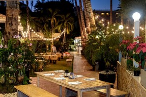 An outdoor dining area with a long wooden table set for two, surrounded by potted plants and string lights. - Lokal Phuket (Patong)