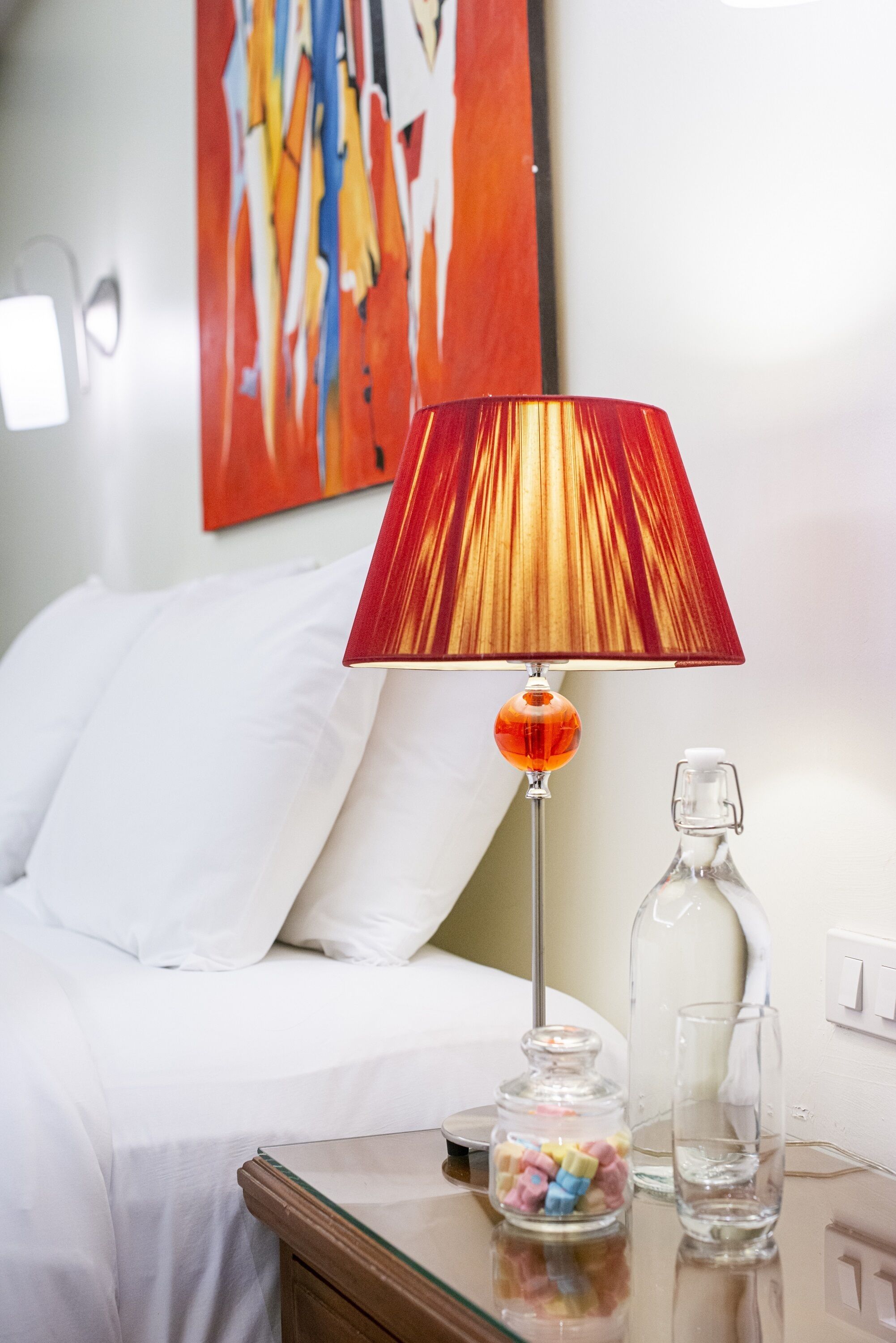 Un lit bien fait, avec du linge de lit blanc, une table de chevet sur laquelle se trouve un bocal en verre rempli de bonbons, un verre d’eau transparent et un abat-jour rouge.