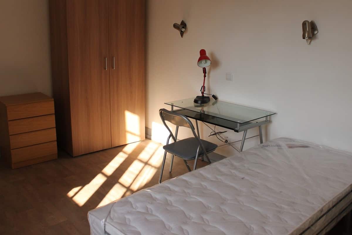 A bedroom with a bed, a desk, a chair, a wardrobe, and a lamp.