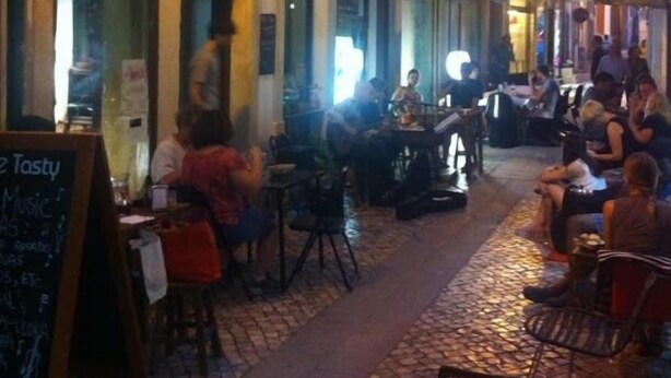 An outdoor café with tables and chairs, a chalkboard menu, and a street view with buildings and people. - Be Coimbra Hostels (Coimbra)