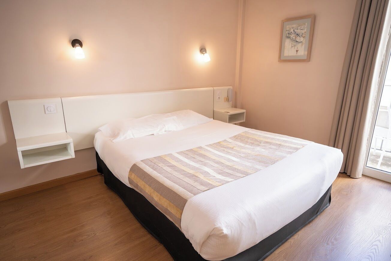 A bedroom with a bed, bedside tables, a nightstand, a wall-mounted shelf, and a window with curtains.