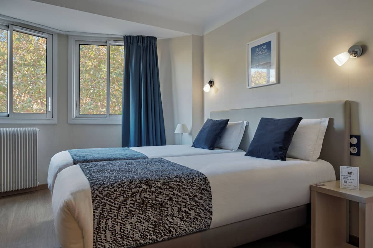 Habitación de hotel con una cama grande, dos almohadas, una mesita de noche, una lámpara y una ventana con cortinas.