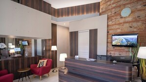 A modern reception area with a wooden reception desk, red chairs, a flat-screen TV, and a large mirror. - The Originals City, Hôtel Mondial, Perpignan (Perpignan)