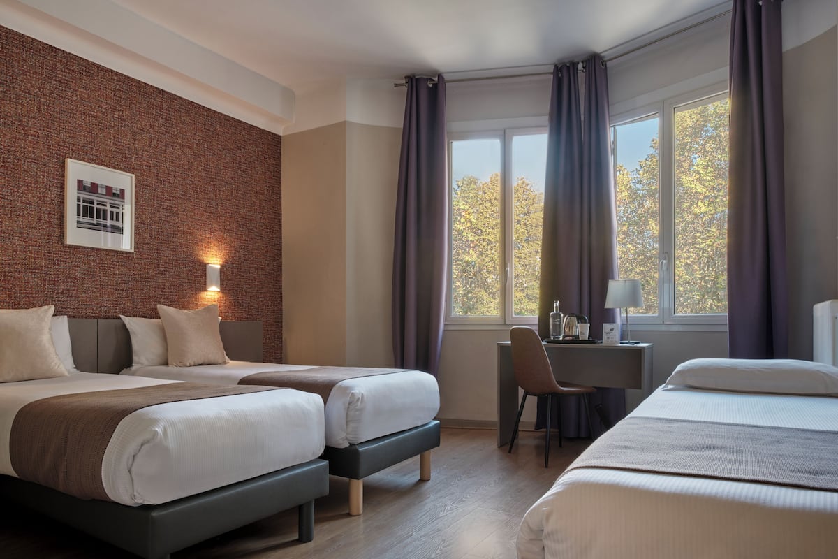 Habitación de hotel con dos camas, un escritorio, una silla, un ventanal con cortinas y una pared de ladrillo con un cuadro enmarcado.