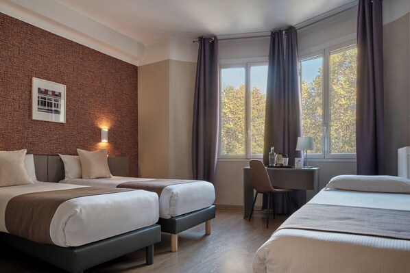 Quarto de hotel com duas camas, uma escrivaninha, uma cadeira, uma janela com cortinas e uma parede de tijolos com um quadro emoldurado.