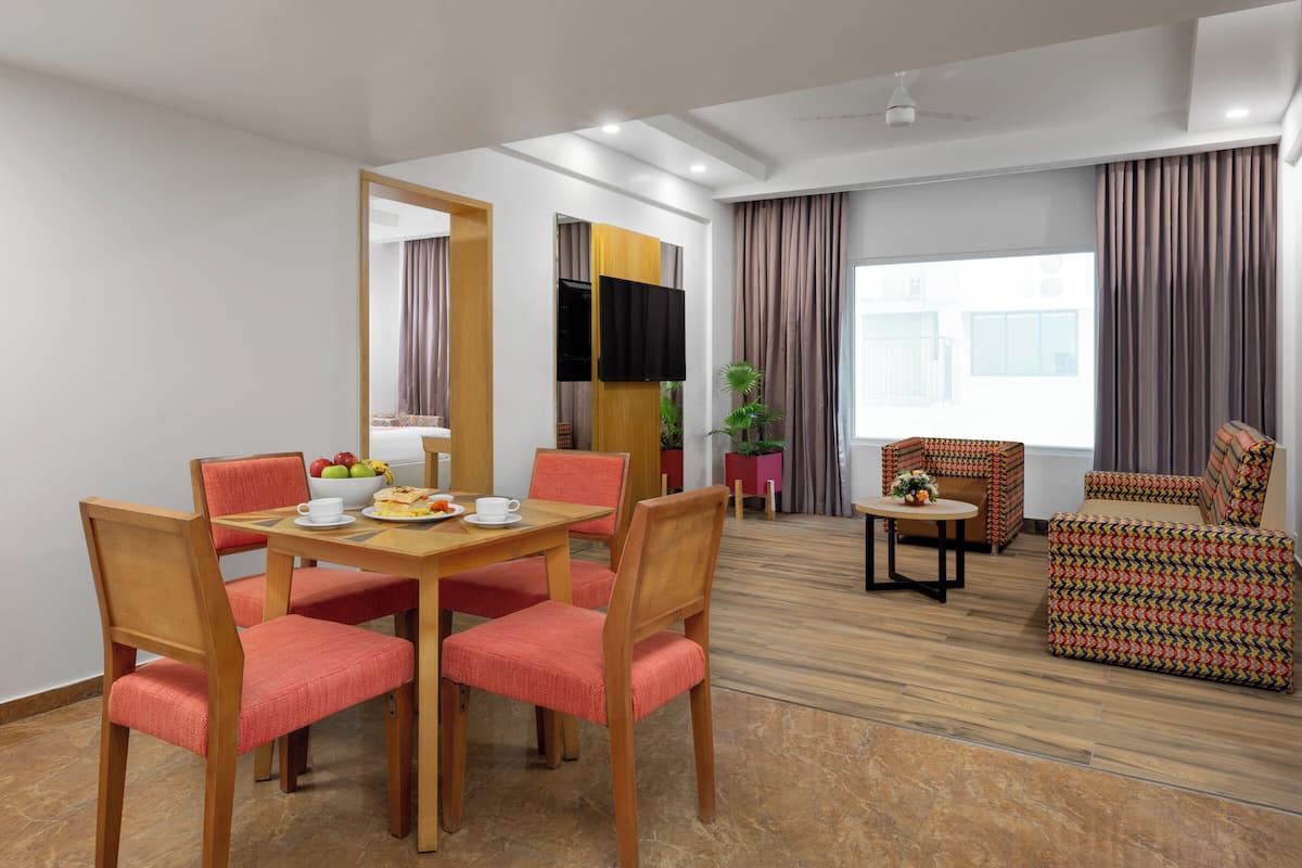 A modern hotel room with a dining area, a sofa set, a television, and a large window with curtains.