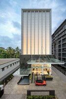 Un edificio moderno con entrada de vidrio, un automóvil rojo estacionado al frente y follaje circundante.