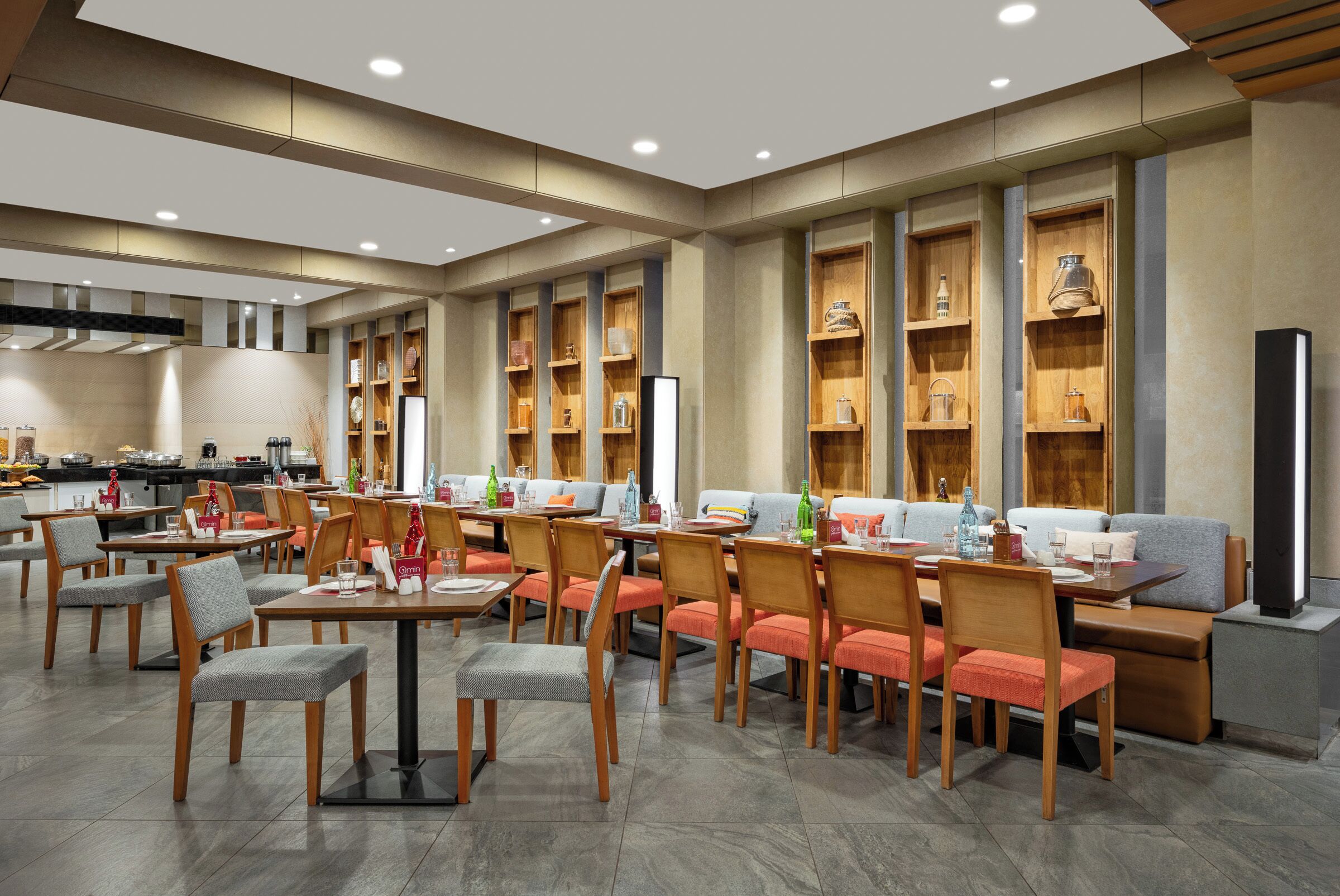 Un área de comedor moderna con varias mesas y sillas, estantes de madera con diversos objetos, y una cocina al fondo.