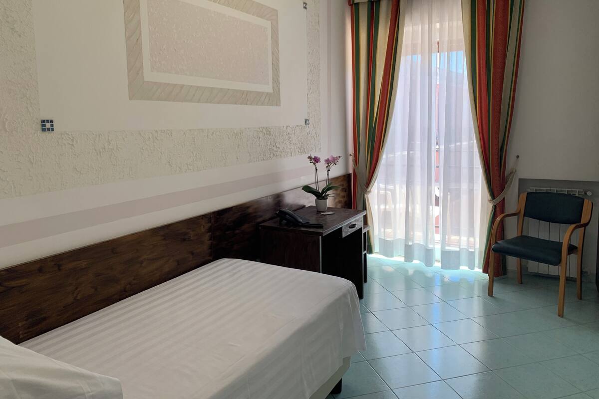 Ein Hotelzimmer mit einem Bett, einem Schreibtisch mit Computer, einem Stuhl und einem fensterbepflanzten Fenster.