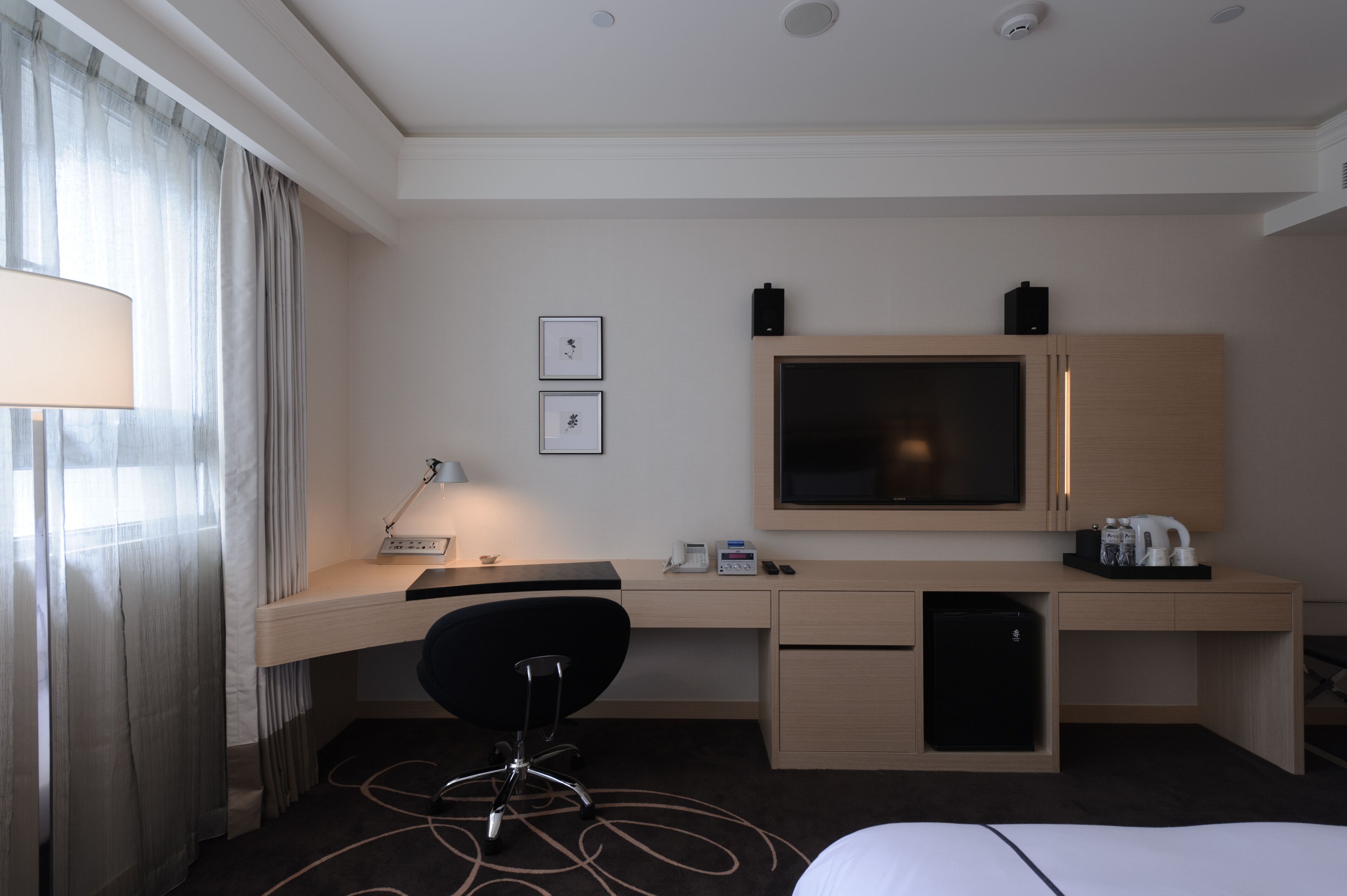 Quarto de hotel com escrivaninha, cadeira, televisão e um pequeno frigorífico.