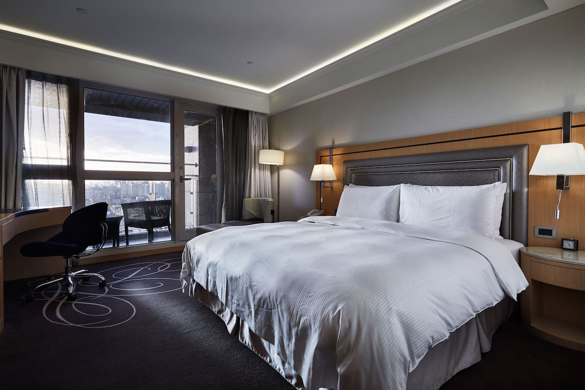 Ein Hotelzimmer mit einem großen Bett, einem Schreibtisch mit Stuhl, einem Fenster mit Vorhängen und Blick auf die Stadt.