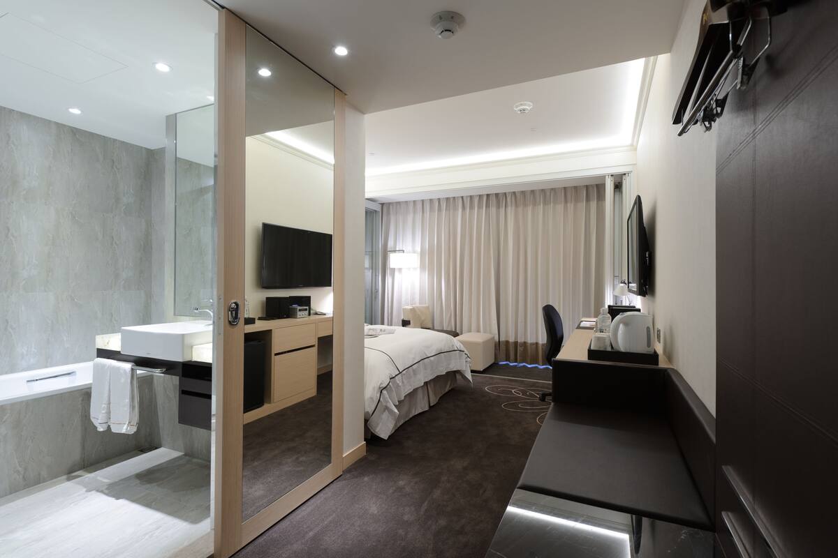 Ein modernes Hotelzimmer mit einem großen Bett, einem eigenen Badezimmer mit Badewanne, einem Schreibtisch mit Stuhl und einem flachbildfernseher, der an der Wand montiert ist.