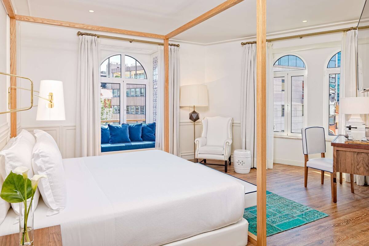 Ruime slaapkamer met een groot bed, een vensterbank met blauwe kussens, een stoel, een bureau en een kleine prullenbak.