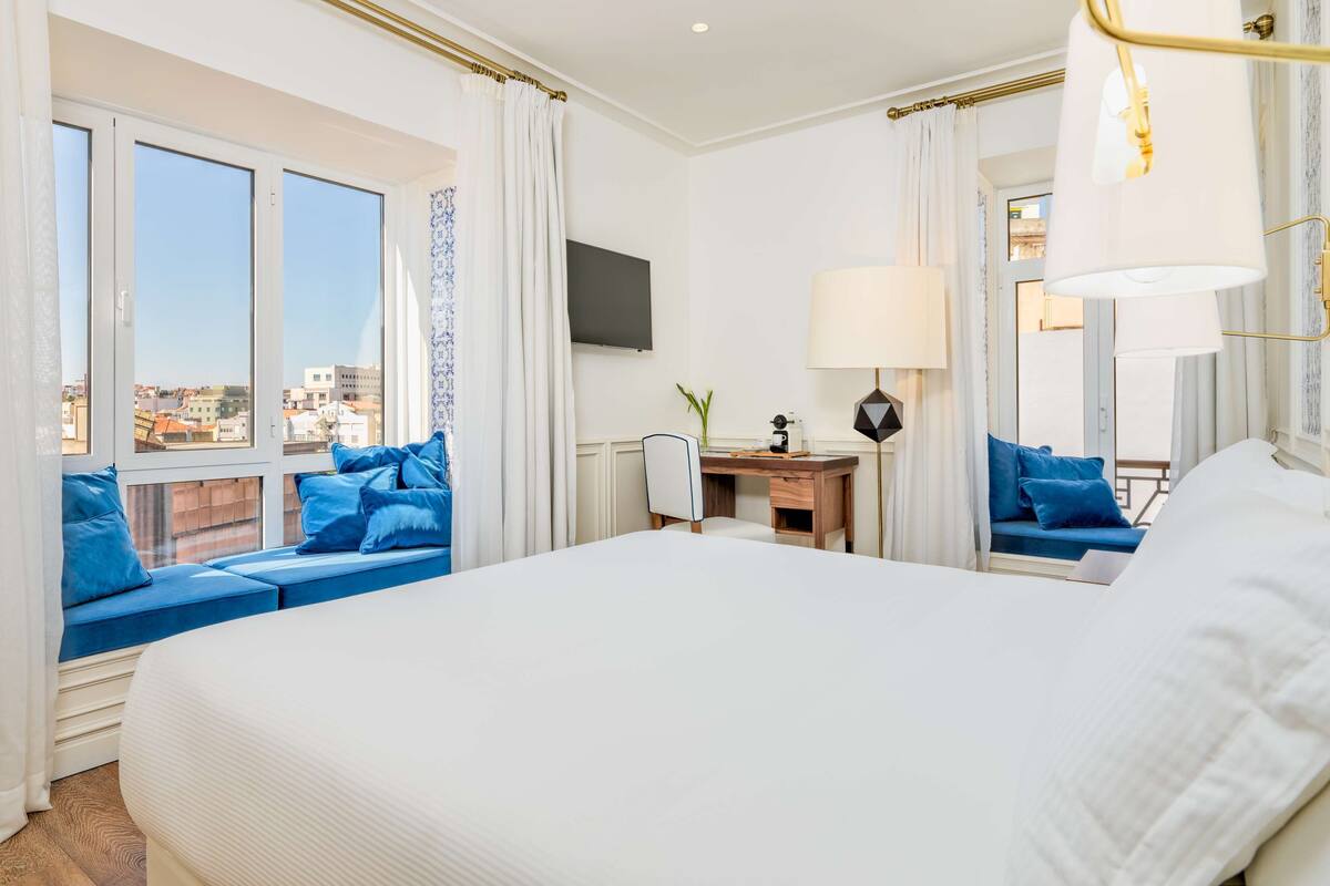 Hotelkamer met een groot bed, een bureau met lamp, een stoel en uitzicht op de stad door grote ramen.