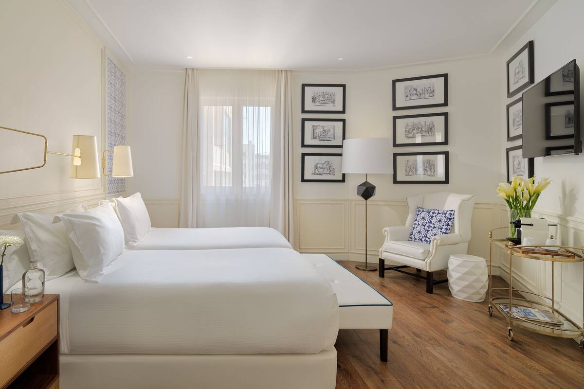 Hotelkamer met twee bedden, een stoel, een bijzettafeltje, een lamp, een vaas met bloemen en ingelijste foto's aan de muur.
