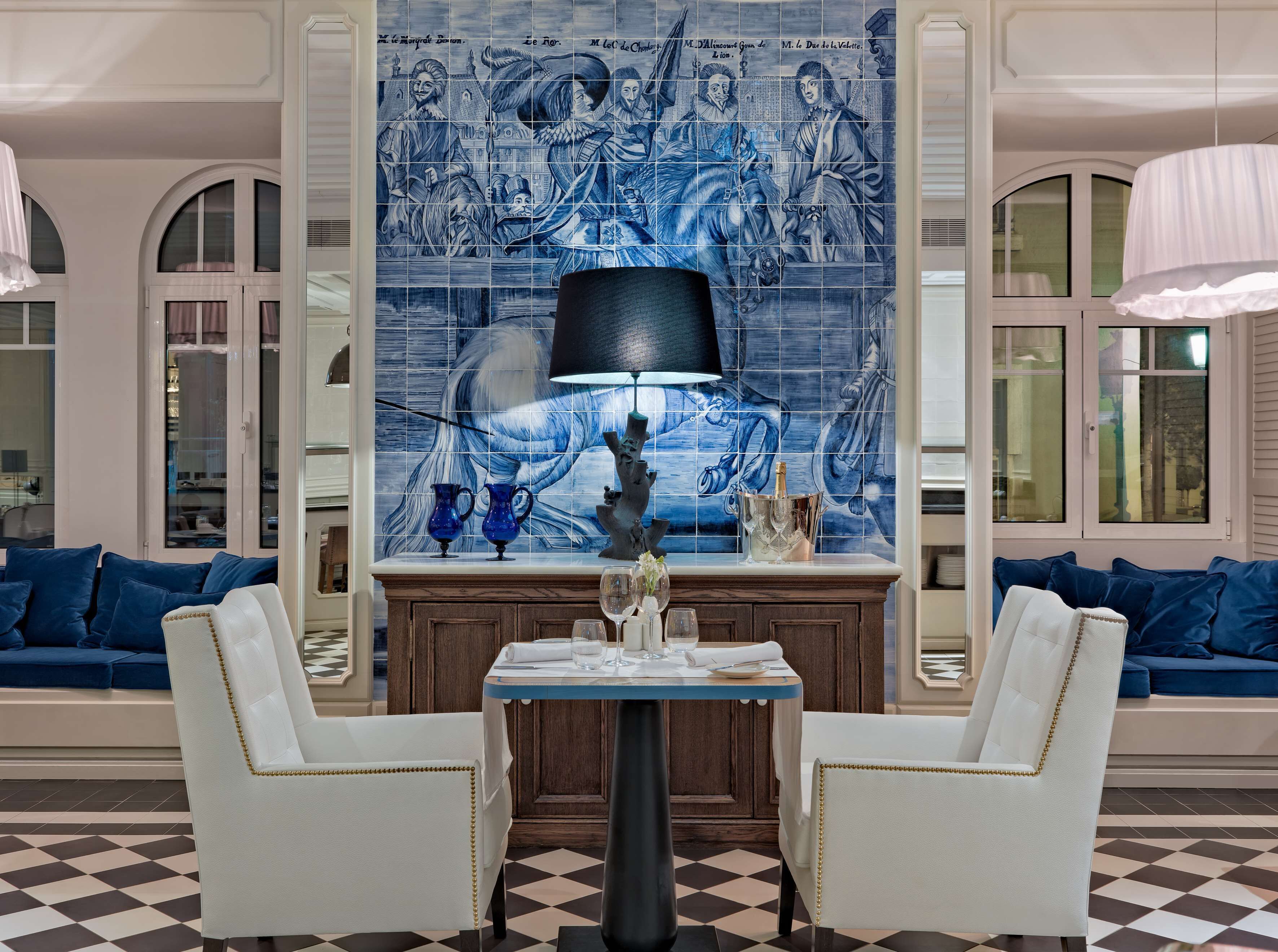 Ein Essbereich mit Schachbrettmuster-Boden, einem Tisch mit Stühlen, einer blauen Wand mit Kacheln und Kunstwerken sowie blauen, gepolsterten Sofas.