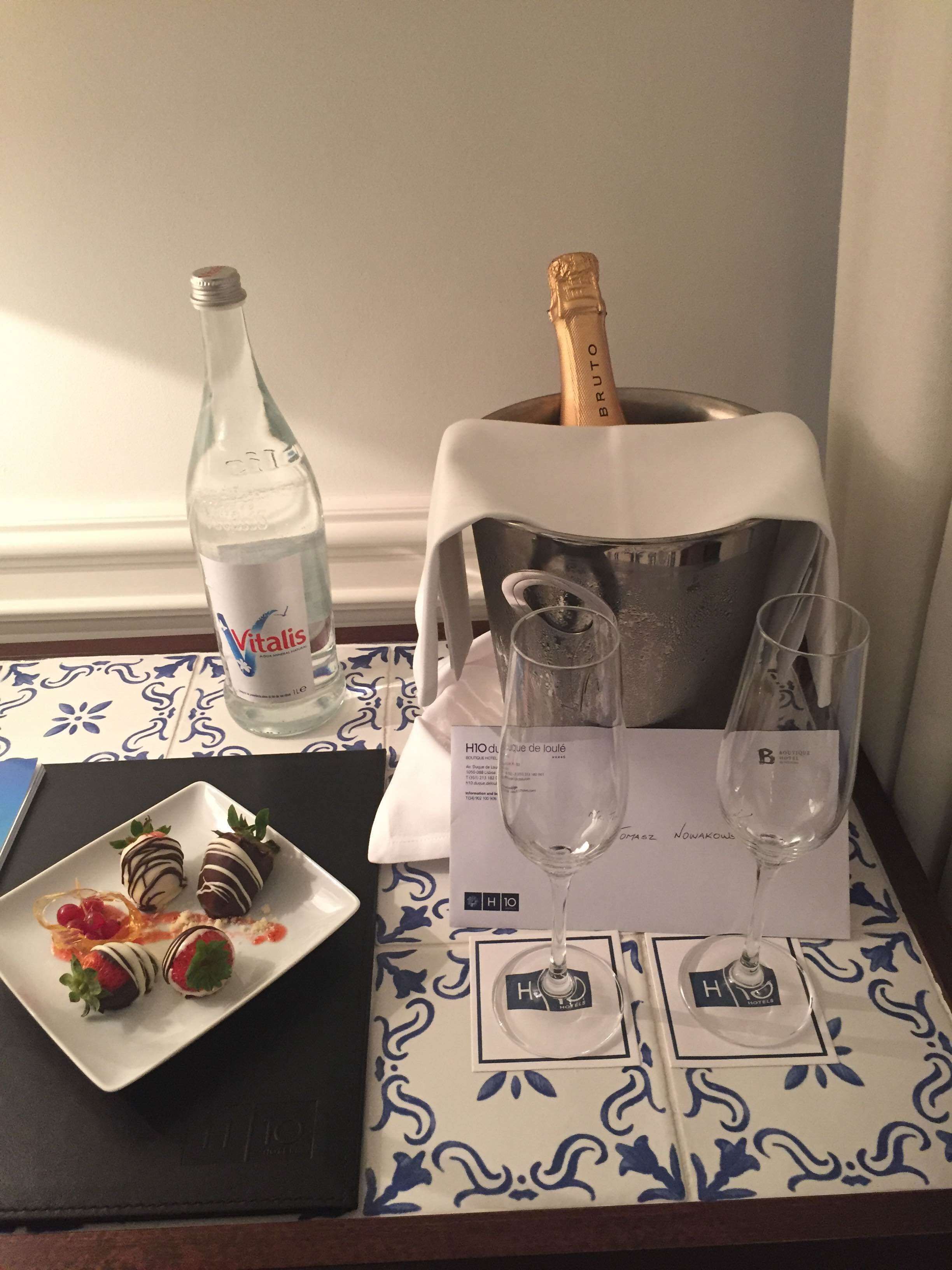 Een tafel met een fles Vitisalys, een fles champagne, twee glazen en een bord met chocoladebedekte aardbeien.
