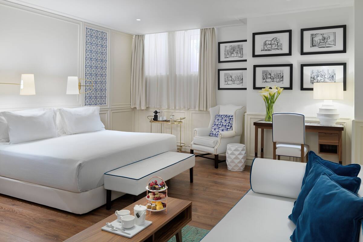 Hotelkamer met een groot bed, een zithoek met een koffietafel, een bureau en een stoel. Er hangen foto's in lijsten aan de muur en er is een raam met gordijnen.