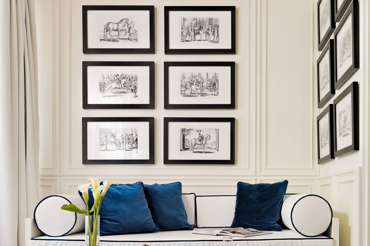 Een kamer met een witte muur, een houten bank met blauwe kussens, een bijzettafeltje met ontbijtartikelen en zwart-wit kunstwerken in lijsten.