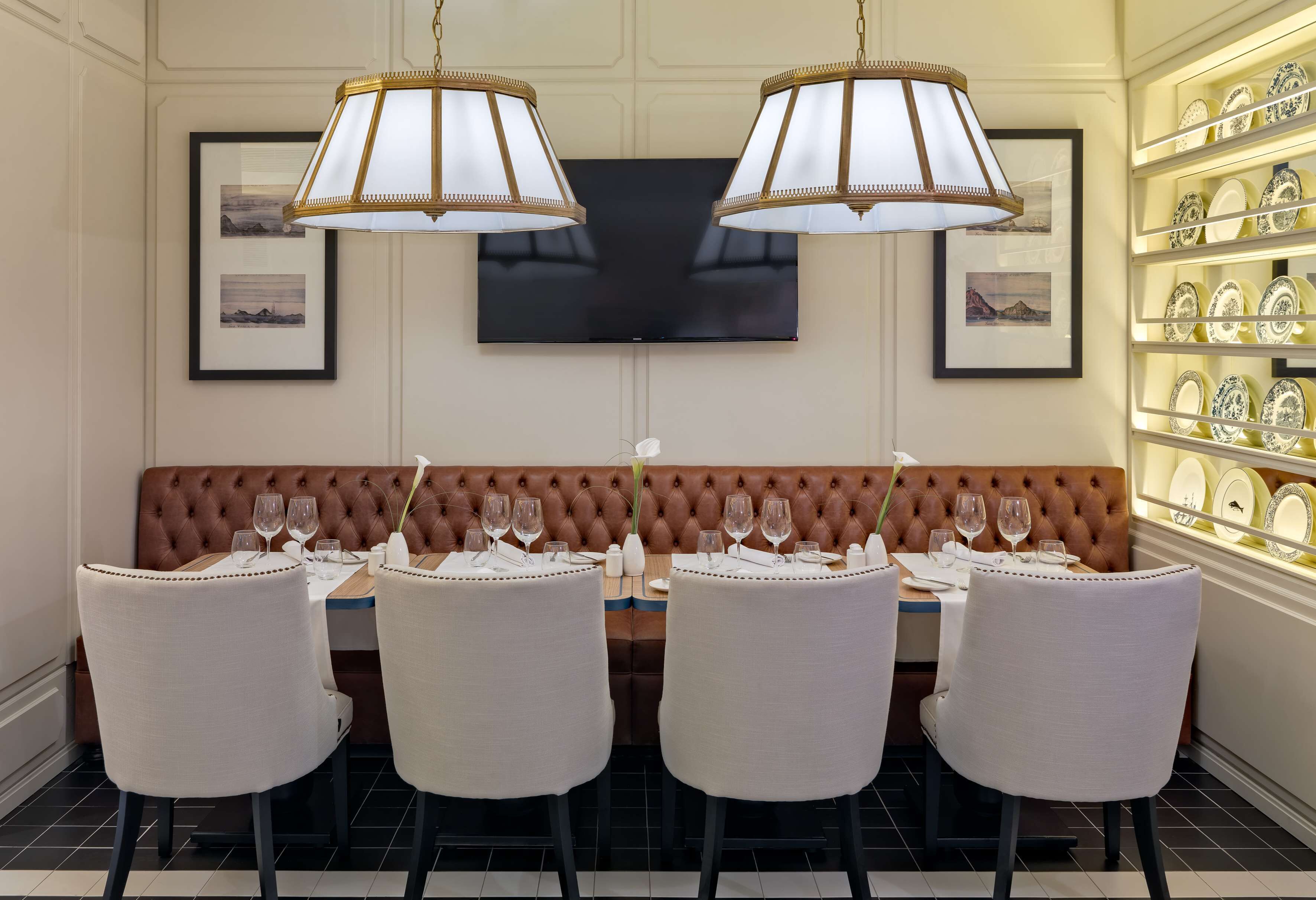 A dining area with a brown upholstered bench, white chairs, a table set with wine glasses, and a television mounted on the wall.