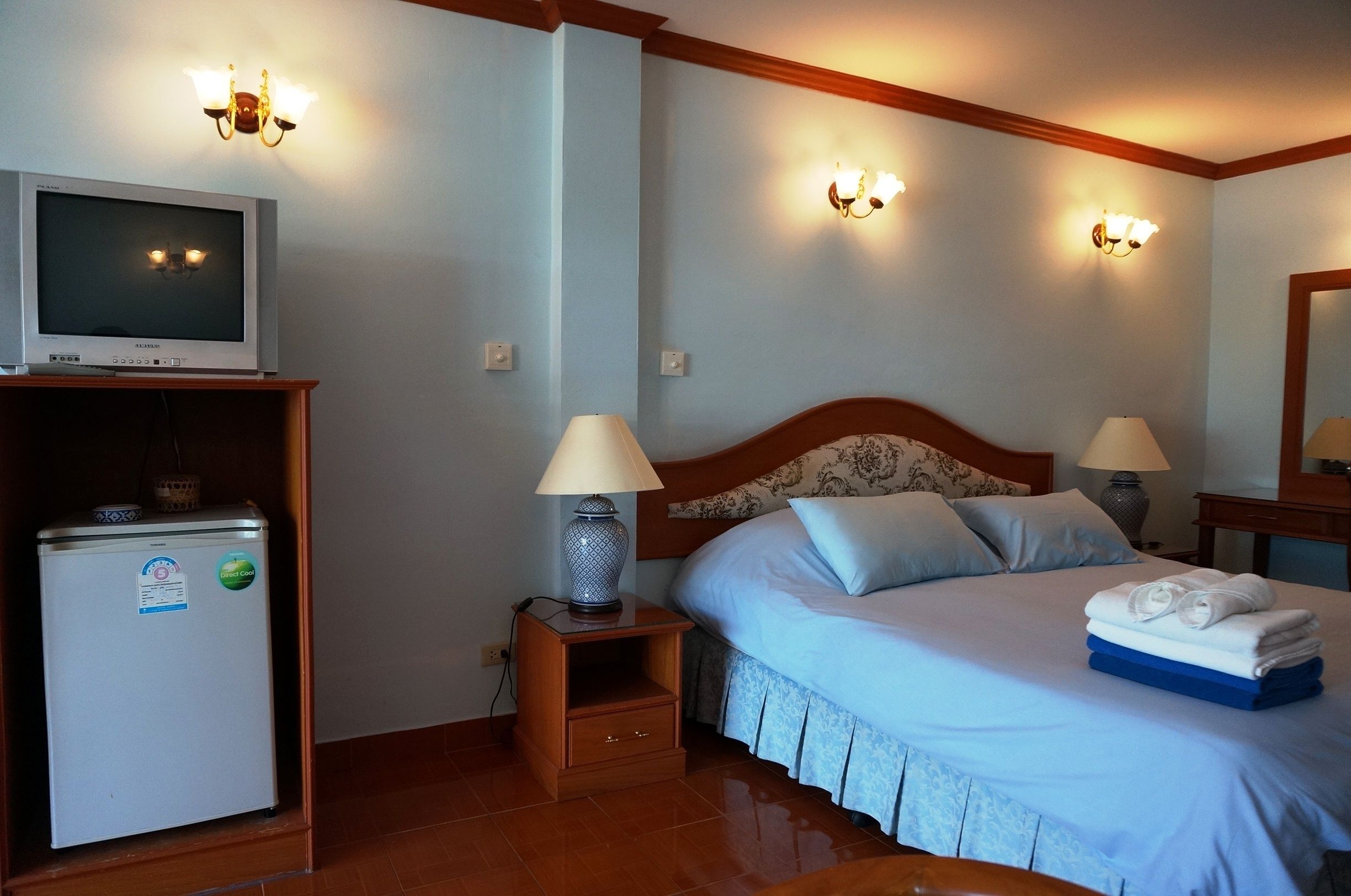 Habitación de hotel con cama, dos lámparas de noche, televisor sobre un mueble bajo, una mesita de noche de madera y un refrigerador.