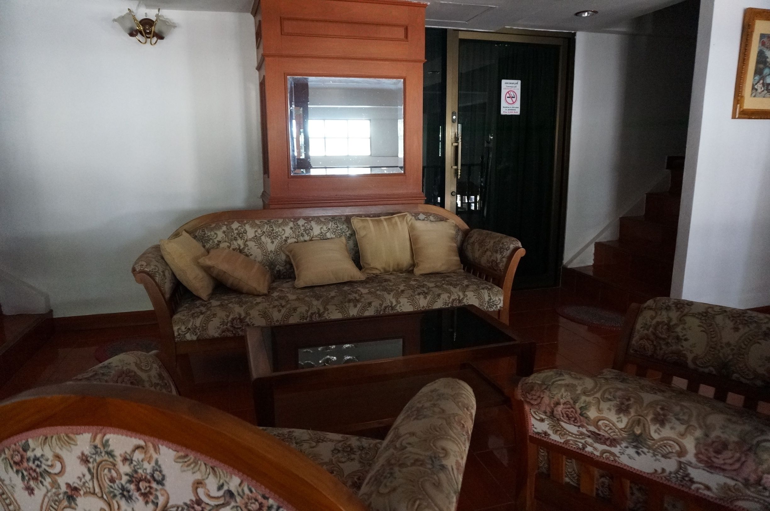 A living room with a sofa, armchairs, and a coffee table. There is a mirror above the sofa and a door with a sign on it.