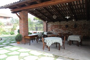 Um pátio externo com paredes de tijolo, mesas e cadeiras de madeira, e uma pérgola com luzes penduradas.