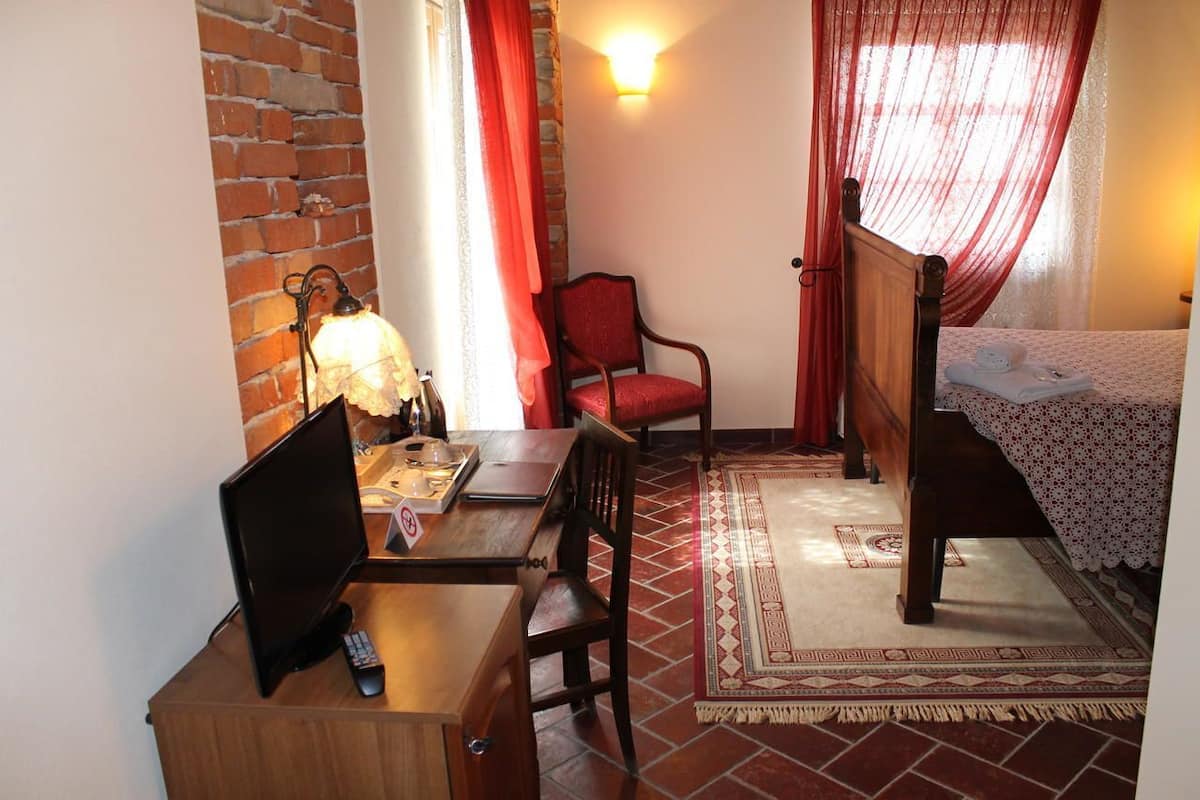 A room with a brick wall, a wooden desk with a computer and remote, a red chair, a bed with a white bedspread, and a patterned rug.