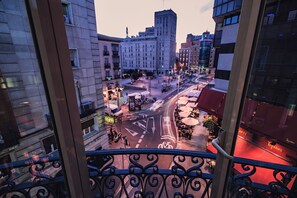 A city street view from a balcony with buildings, a clear sky, and a red-lit storefront. - Hotel Preciados (Madrid)