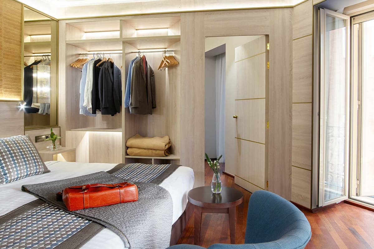 A hotel room with a bed, a bedside table with a vase, a chair, a small table, a closet with hanging clothes, and a sliding glass door to the outside.