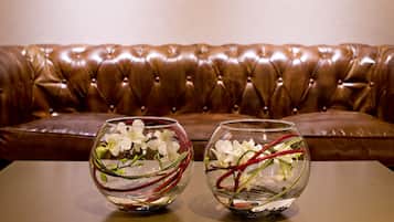 Dois vasos de vidro com flores brancas e caules vermelhos sobre uma mesa, em frente a um sofá de couro marrom.
