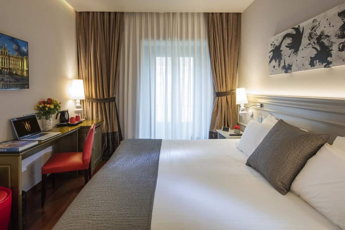 A hotel room with a large bed, a desk with a laptop, a chair, and a red stool. There is a painting on the wall and a window with curtains.